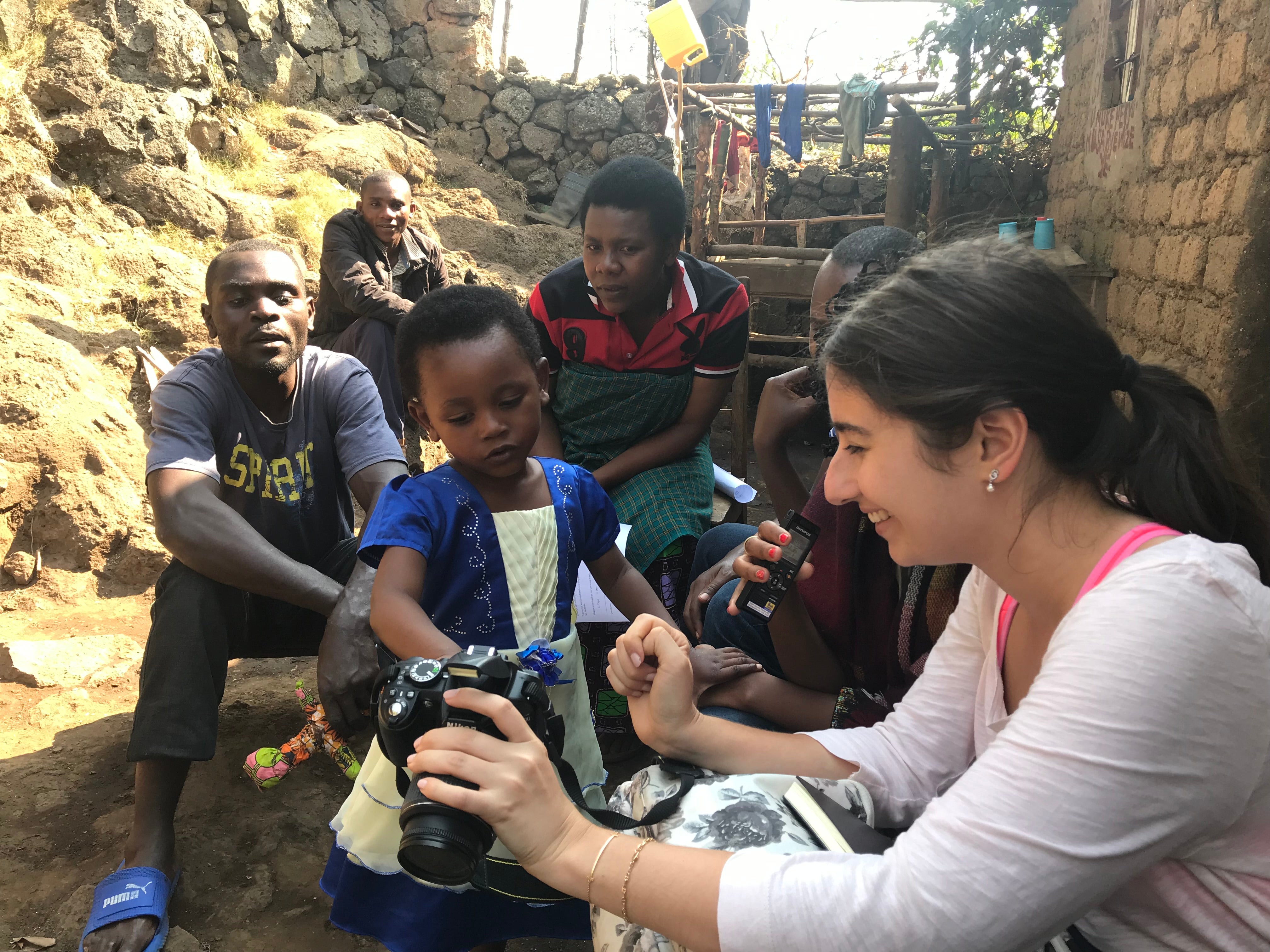 A person shows a camera to a young child while seated outside in a rustic setting with several onlookers in the background.