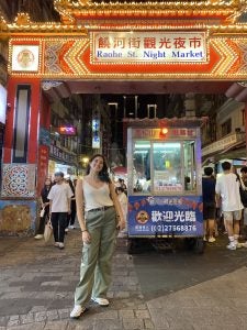Person smiling in front of the brightly lit entrance to Raohe Street Night Market in Taiwan, surrounded by bustling street vendors and visitors.