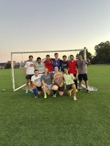 Group of people posing cheerfully in front of a soccer goal on a grassy field.