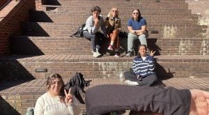 Group of five individuals sitting and posing on brick steps in a sunny outdoor setting, with one person in the foreground making a peace sign.