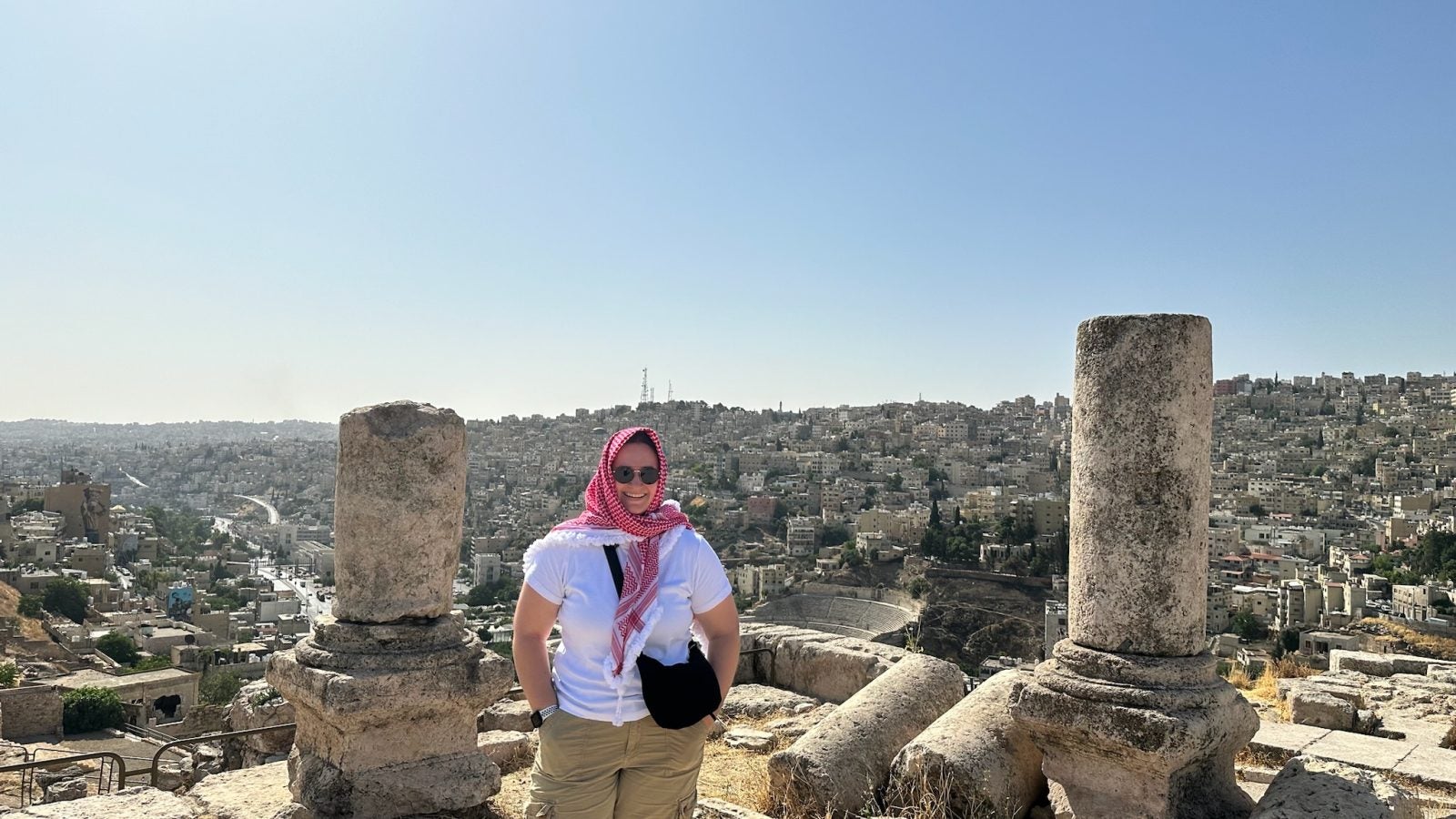 A person wearing a pink scarf and sunglasses stands in front of ancient column ruins with a panoramic view of the cityscape in the background.