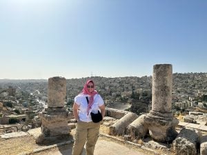 A person wearing a pink scarf and sunglasses stands in front of ancient column ruins with a panoramic view of the cityscape in the background.