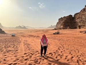 A person stands on the sandy terrain of Wadi Rum at sunset, flanked by large rock formations with vehicles parked in the background.