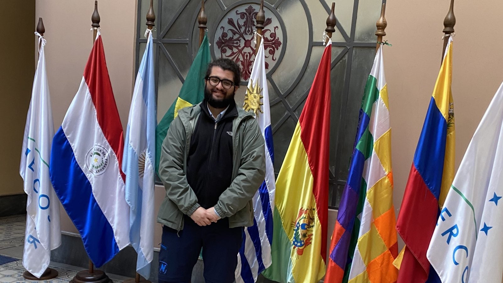 A person smiling and standing in front of a collection of international flags in a tiled hallway.