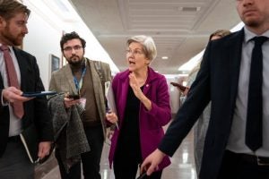 Senator Elizabeth Warren walking and talking with reporters in a corridor.