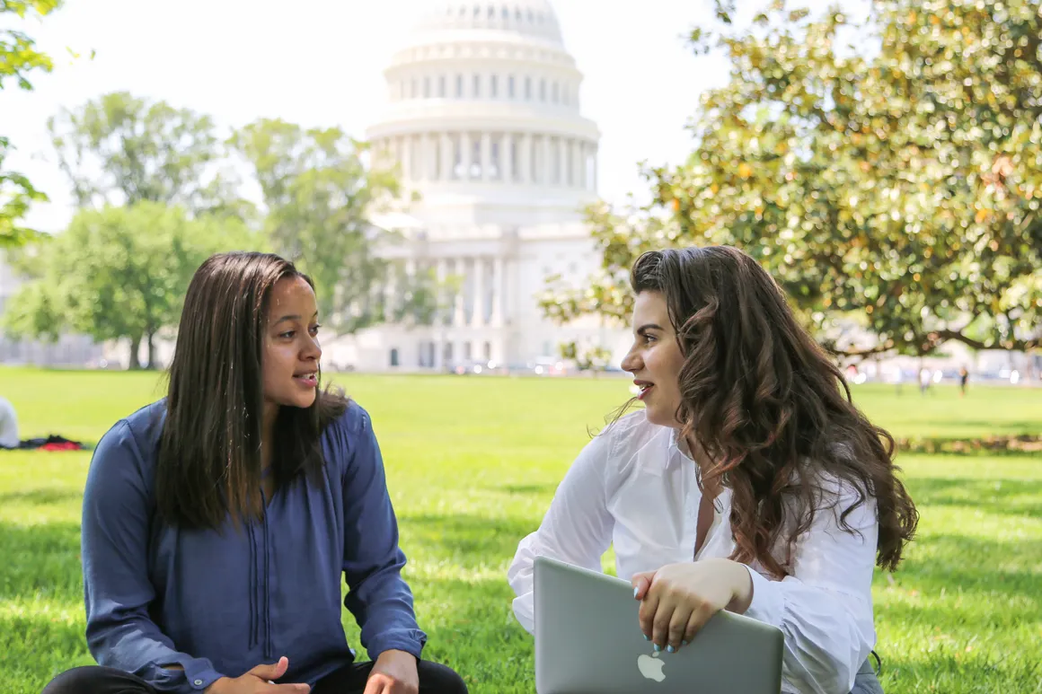 Two students sitting on the lawn talking, with computers in their laps, in front of the U.S. Capitol building