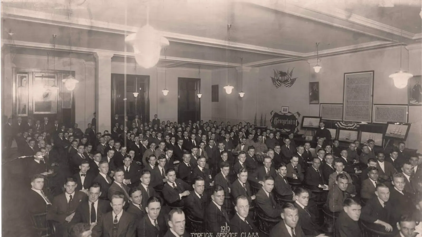 Archival black and white photograph of the first class of the School of Foreign Service in 1919. A large group of around 100 young men in suits sit in a classroom.