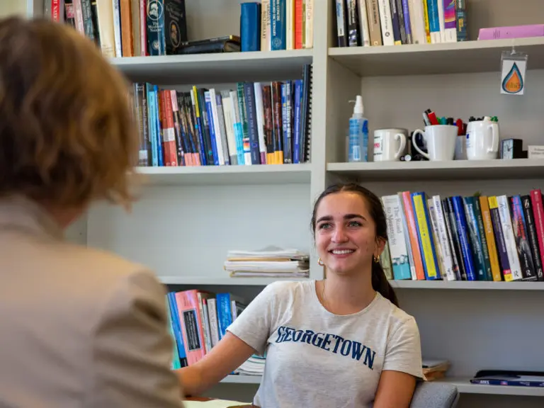 Georgetown student speaking to a woman who is facing the student. behind her a bookshelf filled with books.