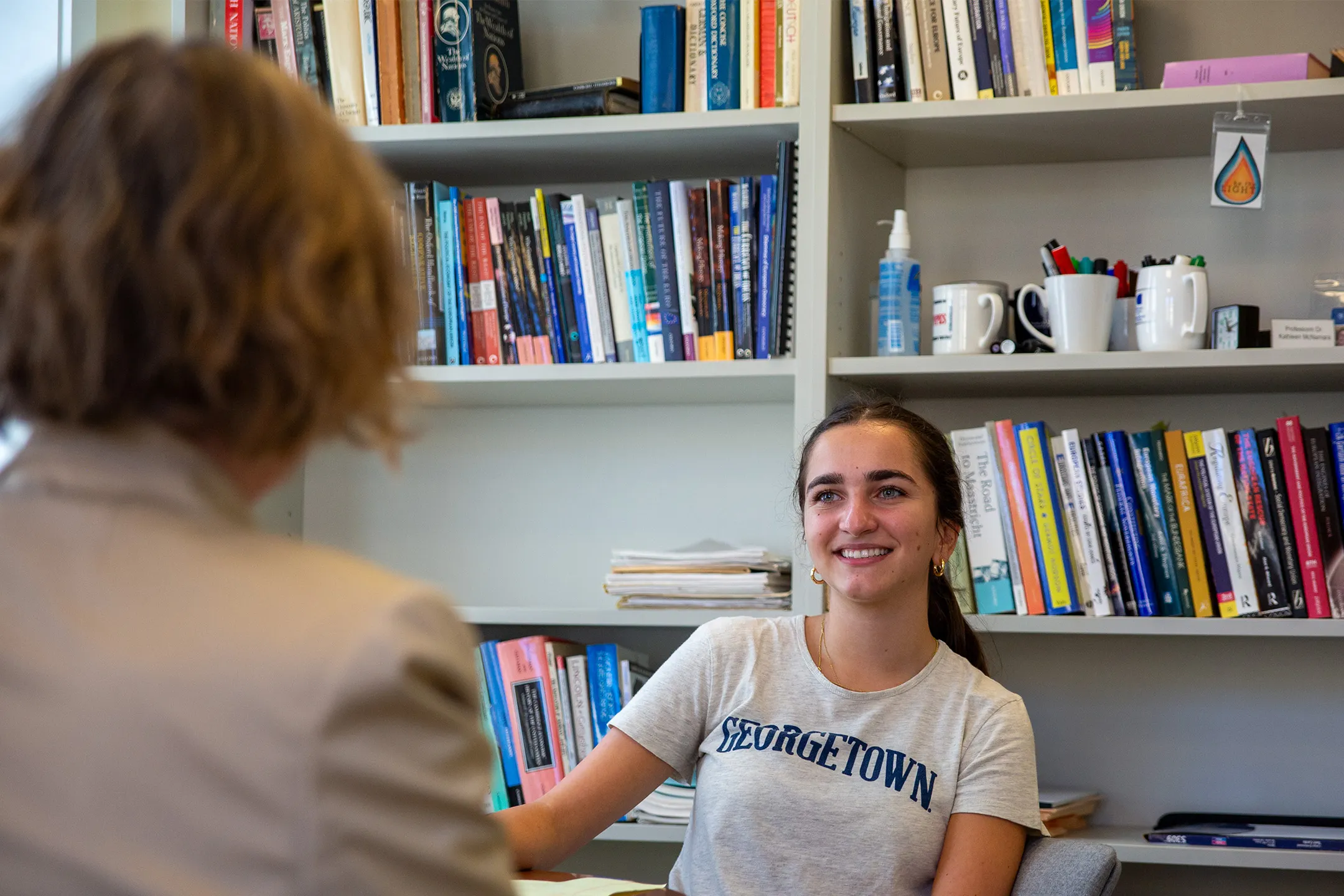 Georgetown student speaking to a woman who is facing the student. behind her a bookshelf filled with books.