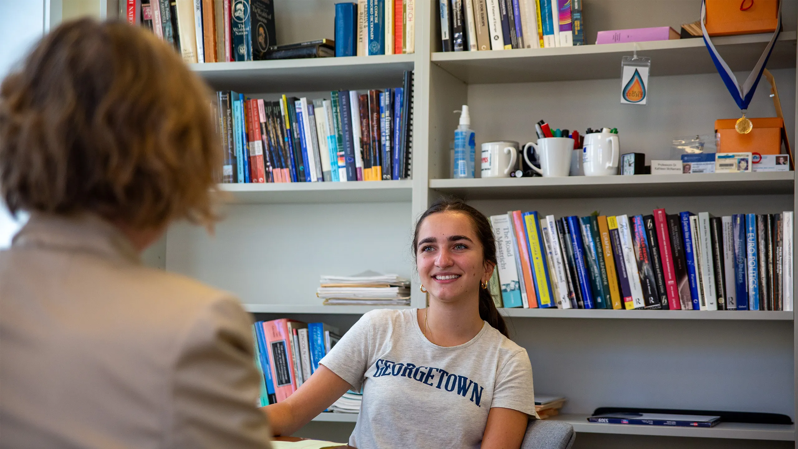 Georgetown student speaking to a woman who is facing the student. behind her a bookshelf filled with books.