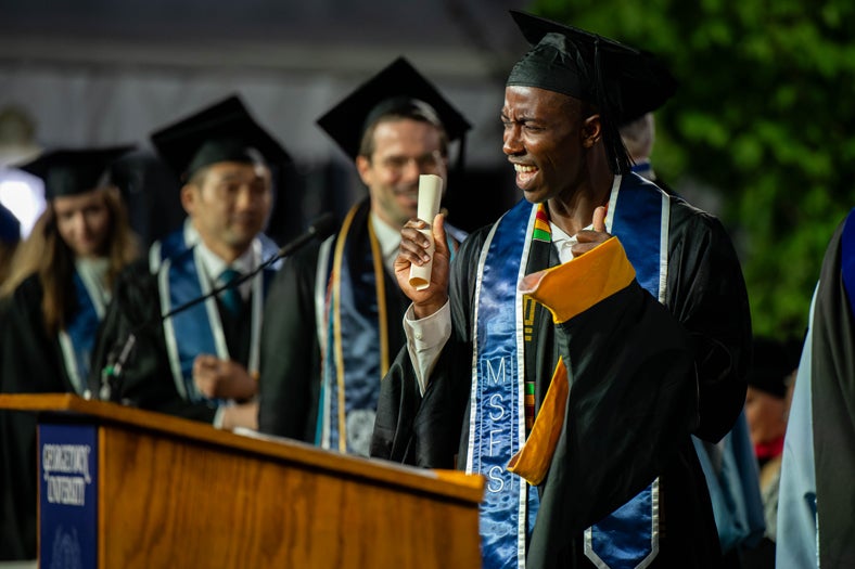 Student wearing a cap and gown in the commencement stage holding his diploma in one hand, and showing a thumbs up in his other hand. He is cheering. Behind him, out of focus are three more students.