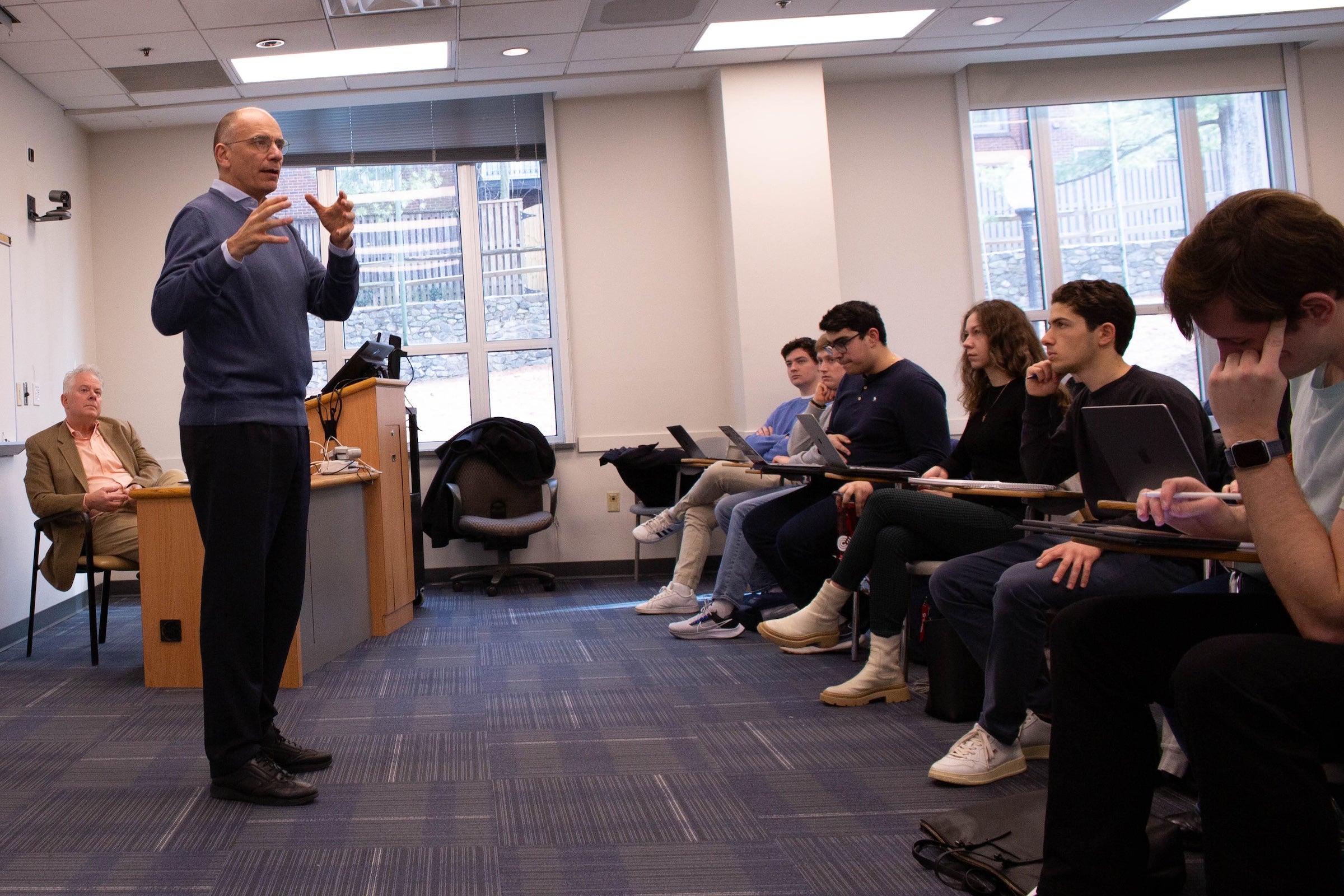 Enrico Letta gestures while standing in a classroom in front of students with Professor Jeffrey Anderson sitting in the background.