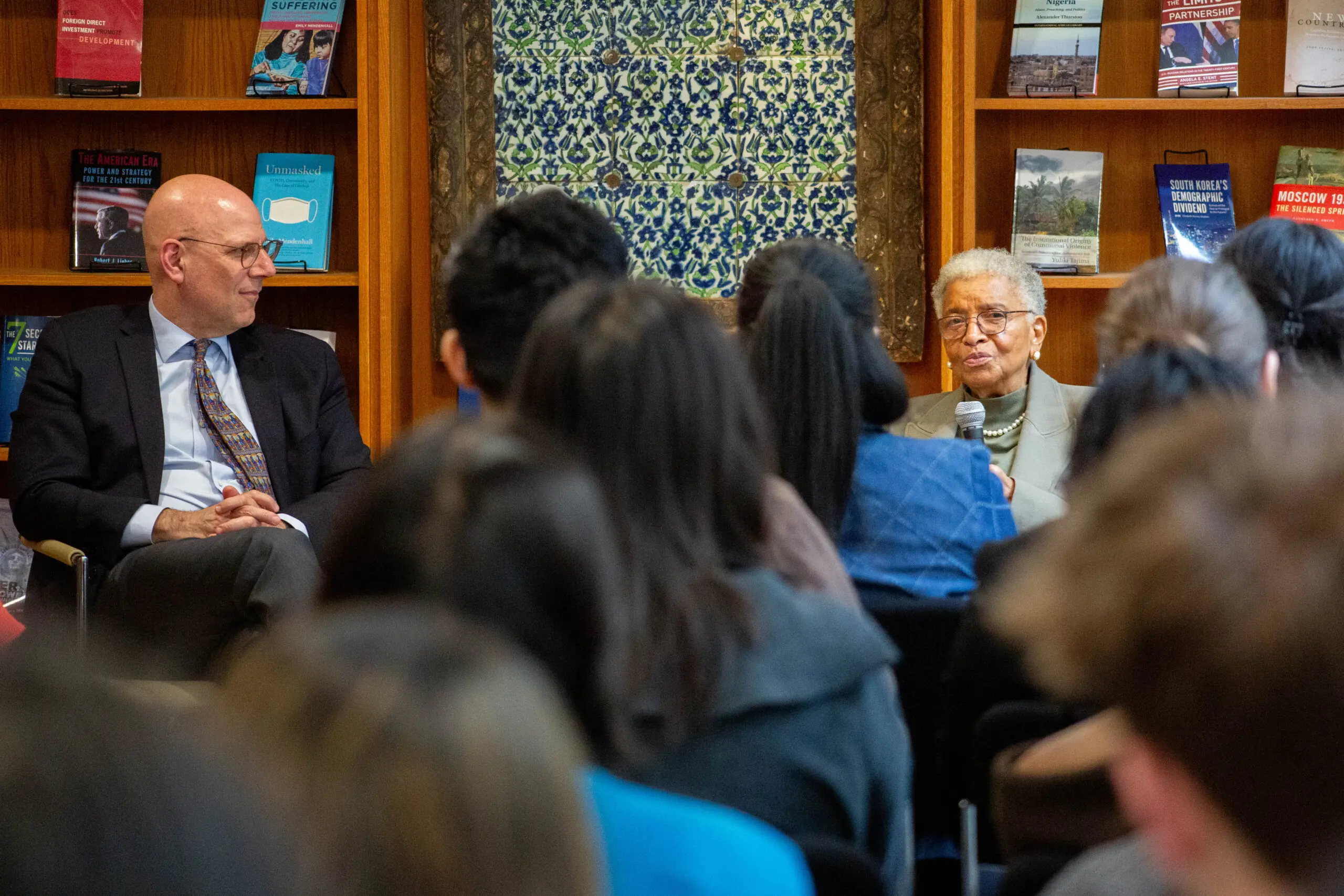 A man and woman speak to an audience during an educational event, with various books displayed in the background.