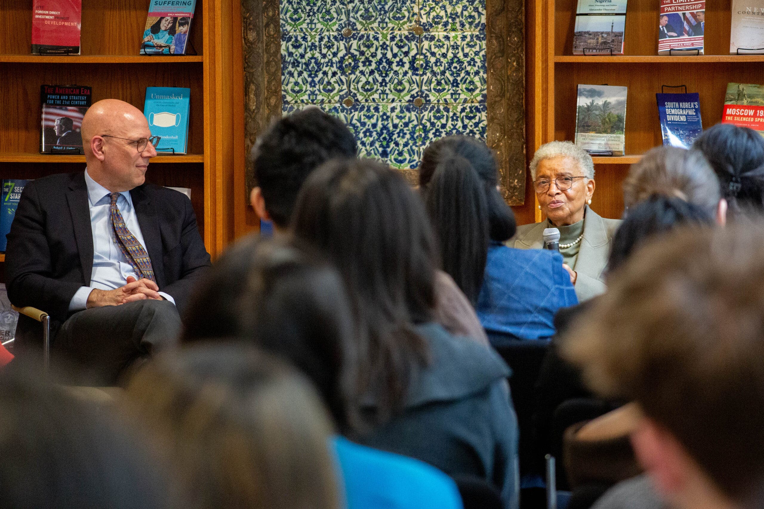 A man and woman speak to an audience during an educational event, with various books displayed in the background.