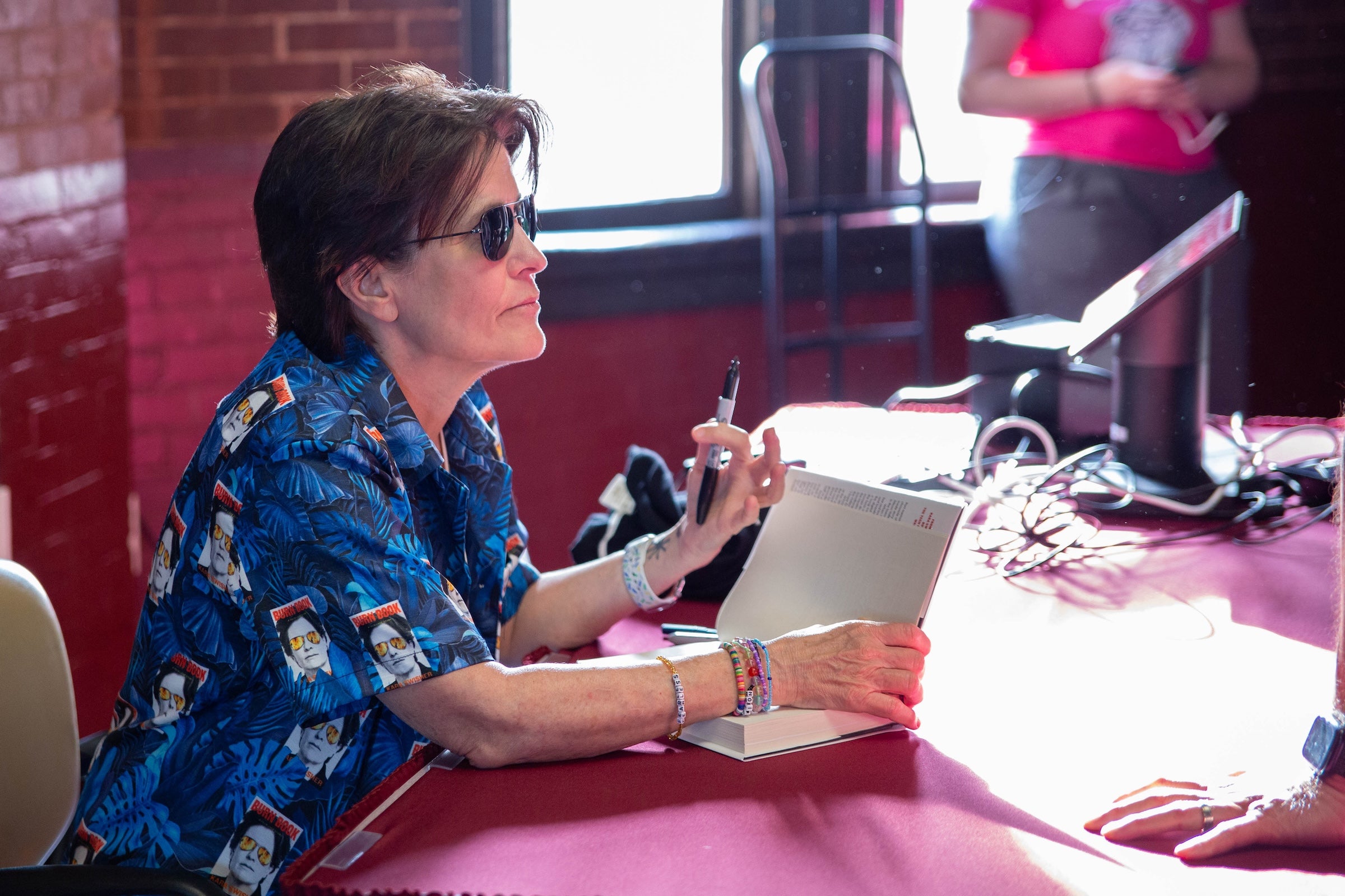 Swisher wearing sunglasses sits at a table signing a copy of her book.