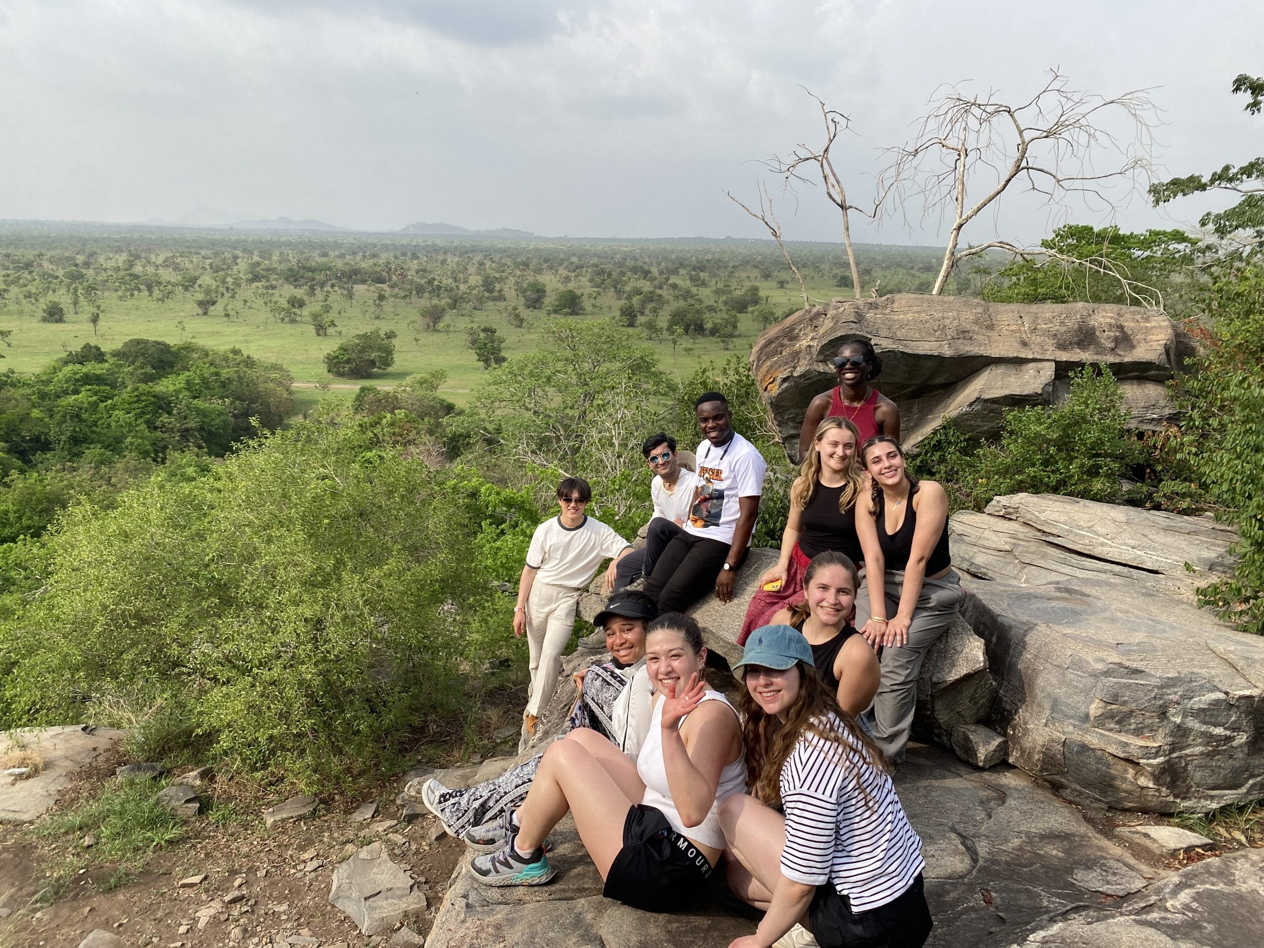 Ten college students in tee shirts, tank tops, and shorts sit on rocks in front of a green field dotted with small green trees.