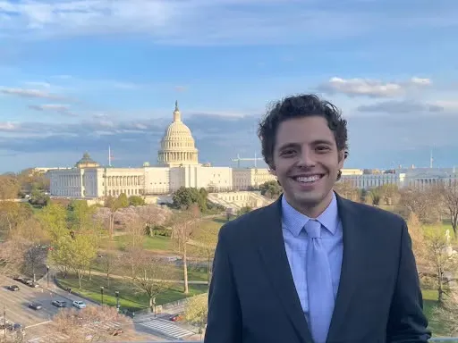 Ali-Caccamo wearing a suit stands on a rooftop with the U.S. Capitol building behind him.