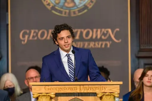 Adrian Ali-Caccamo stands at a podium in Gaston Hall in a suit.