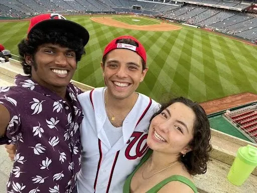 Wearing a Washington Nationals shirt and hat, Adrian Ali-Caccamo stands with his brother Subin and sister Juliet in Nats Park with the field in the background.