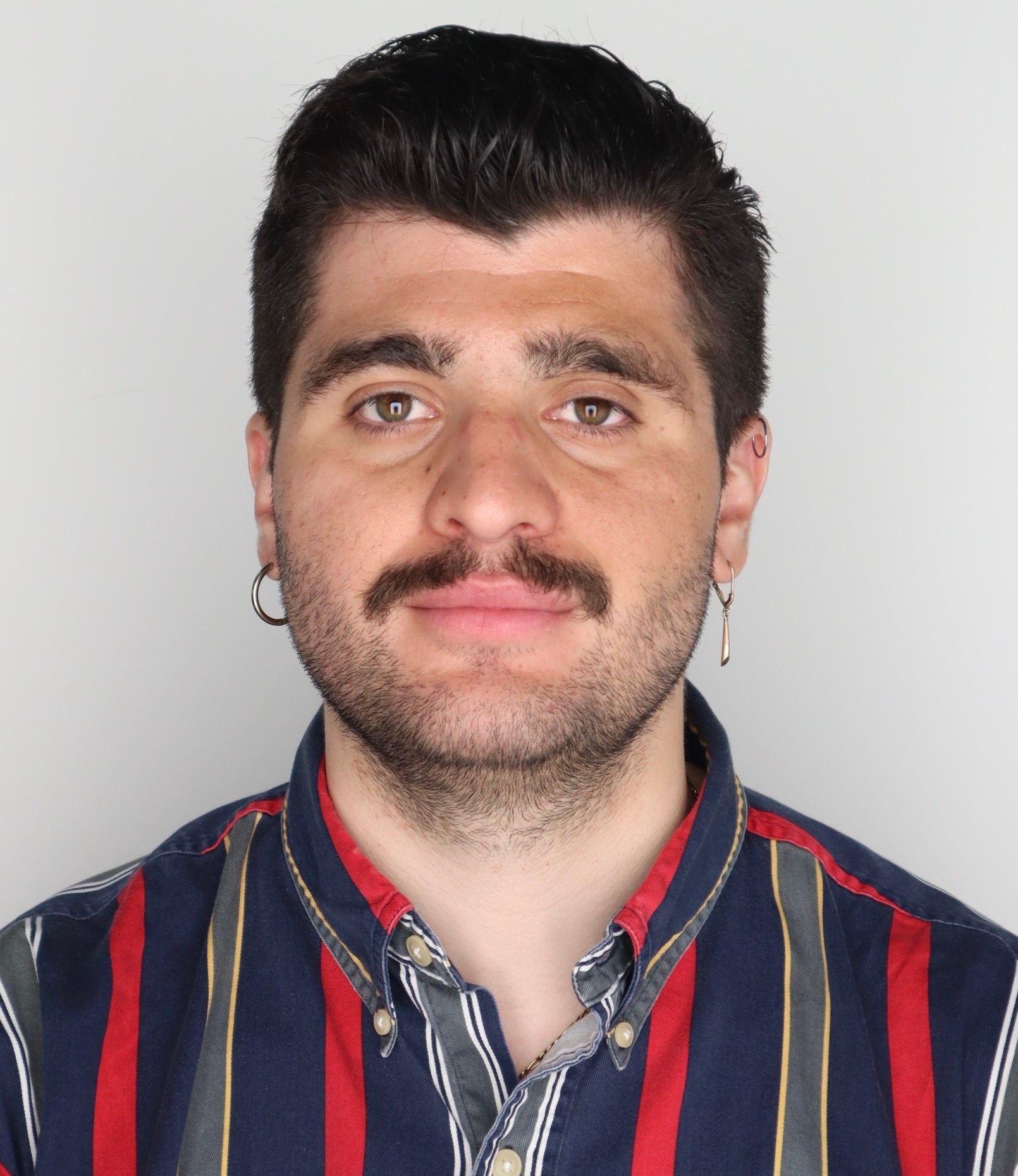 Portrait of a person with a styled haircut and a short beard, wearing a striped shirt and hoop earrings, smiling against a plain light background.