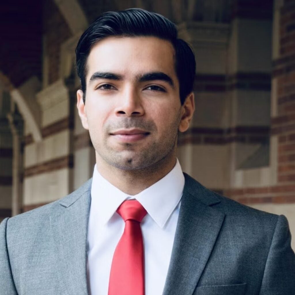 Ali Unar wearing a gray blazer, white, button-down shirt, and a red tie.