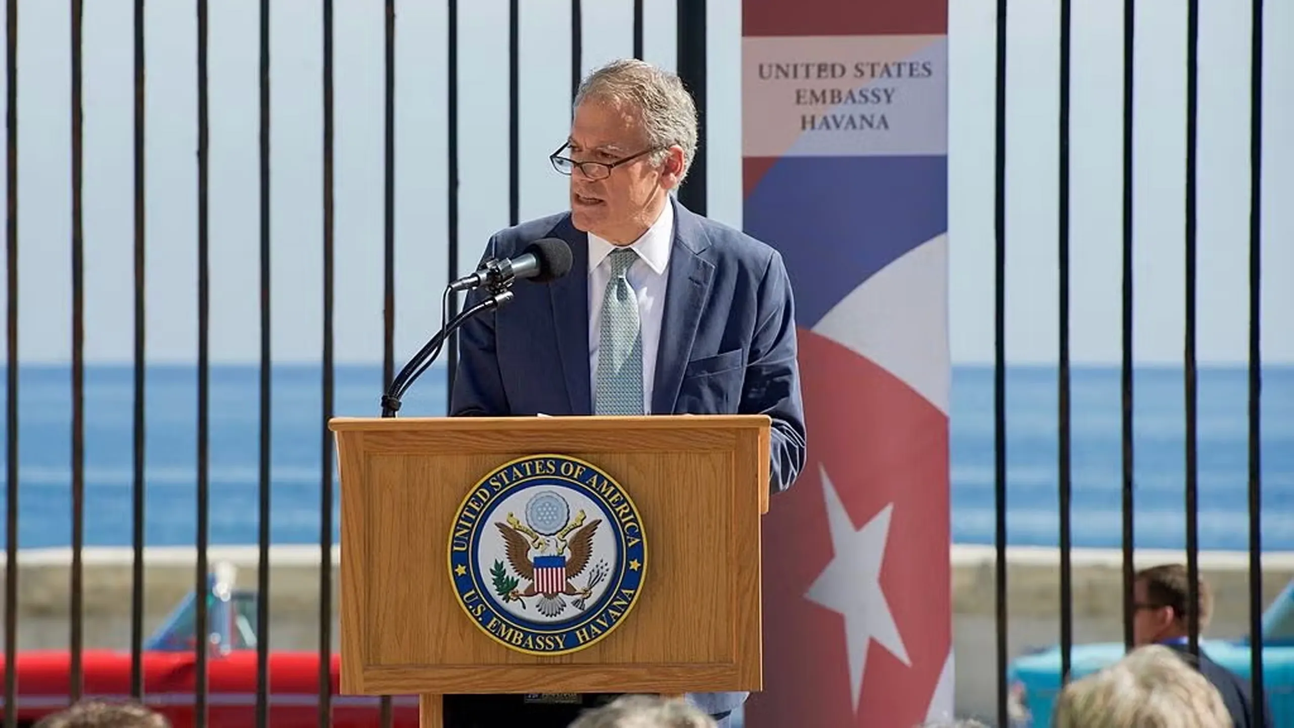 Ambassador Jeffrey DeLaurentis speaking on a United States of America U.S Embassy Havana podium, outside on a sunny day. Behind him, the ocean.