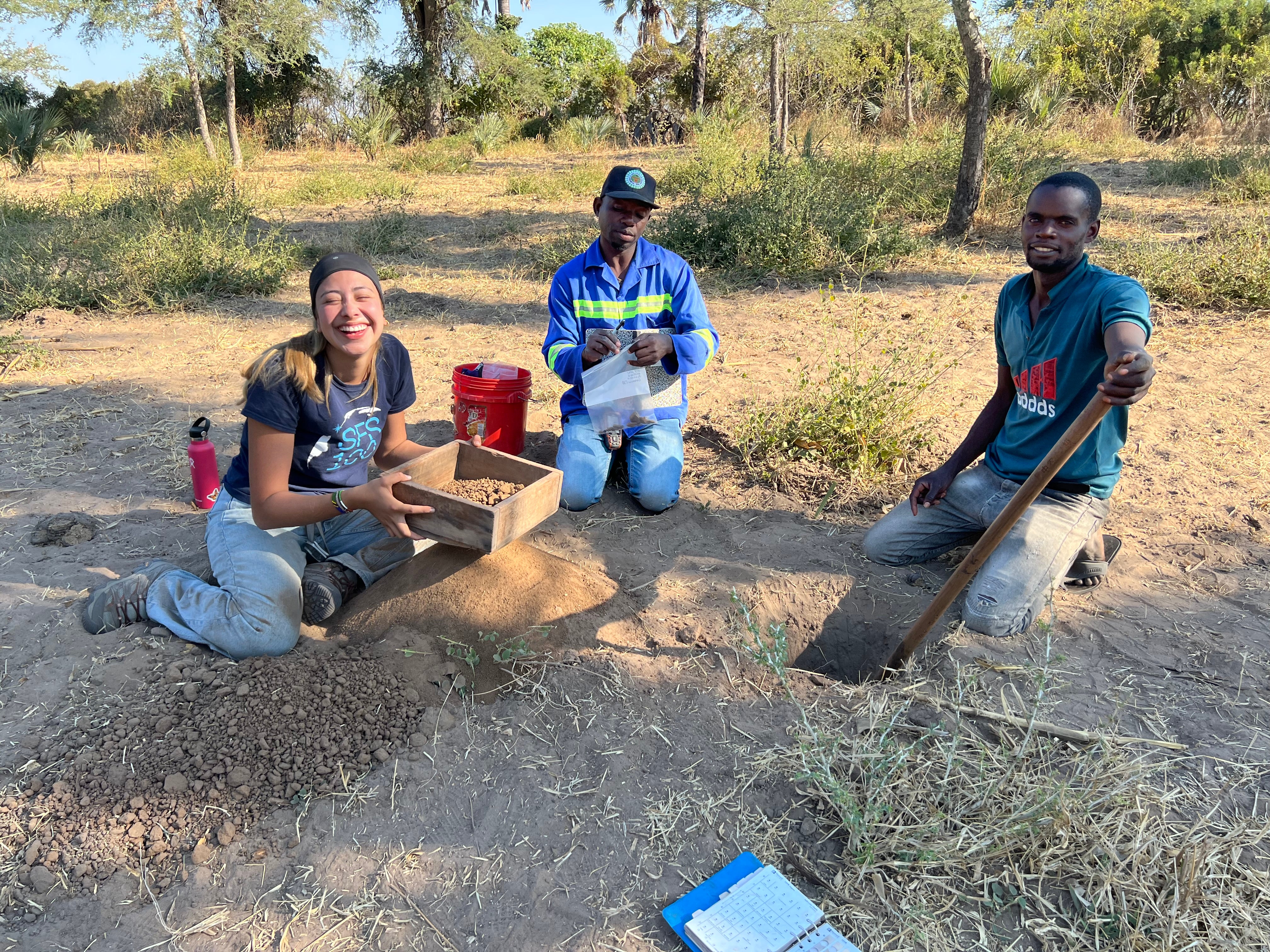 Andrea Nucete kneels on the dirt next to a small hole with two Zambian men with shovels.