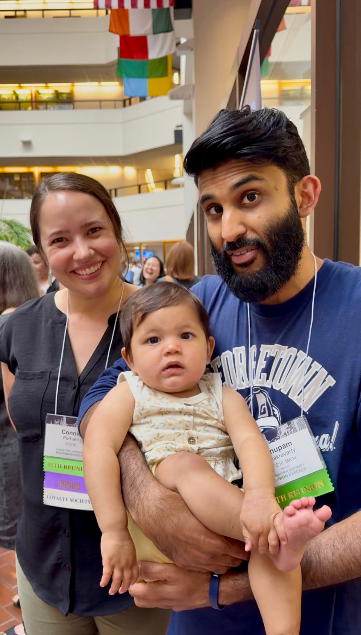 Two adults and a baby at a conference, wearing nametags, smiling in an indoor atrium with flags hanging overhead.