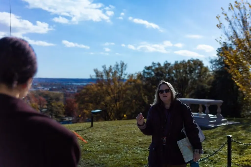 Two individuals are having a conversation outdoors on a sunny day with a scenic overlook featuring a distant cityscape in the background.