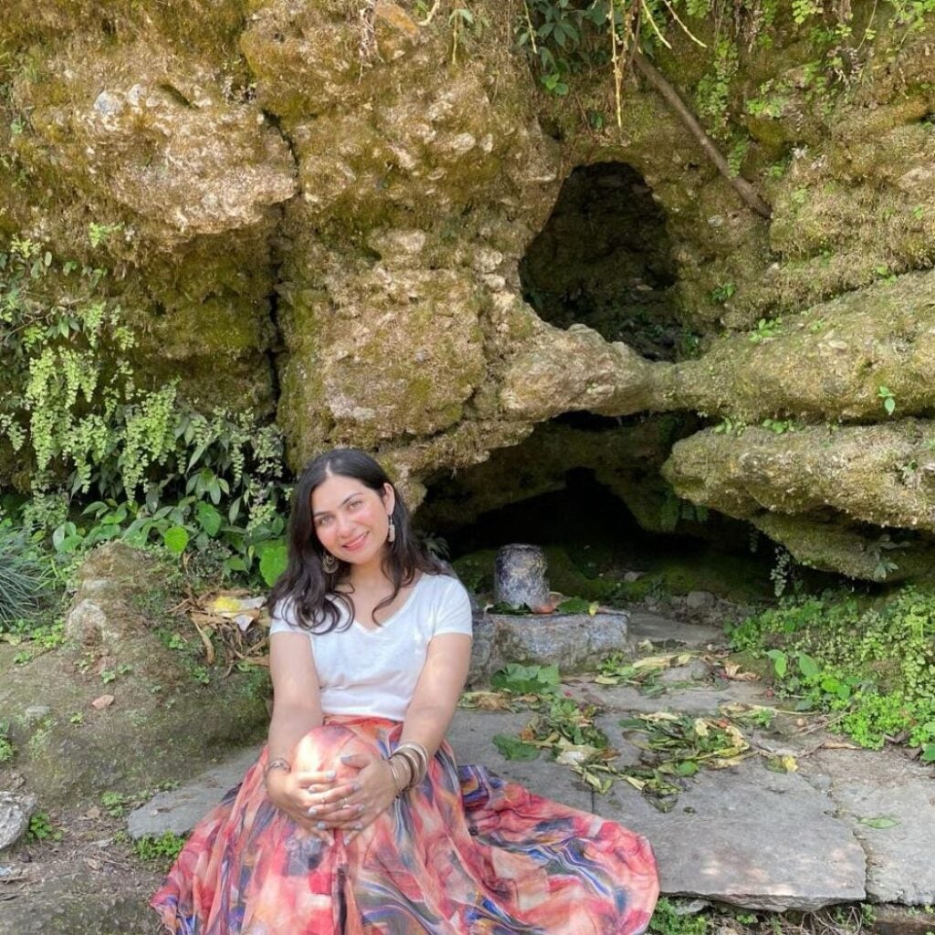 Arshia Chand wearing a multicolored skirt and white blouse, sitting on the ground in front of a cave.