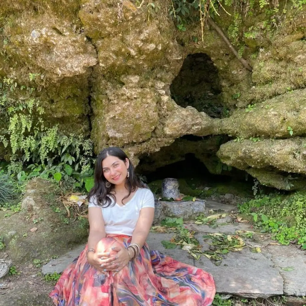 Arshia Chand wearing a multicolored skirt and white blouse, sitting on the ground in front of a cave.