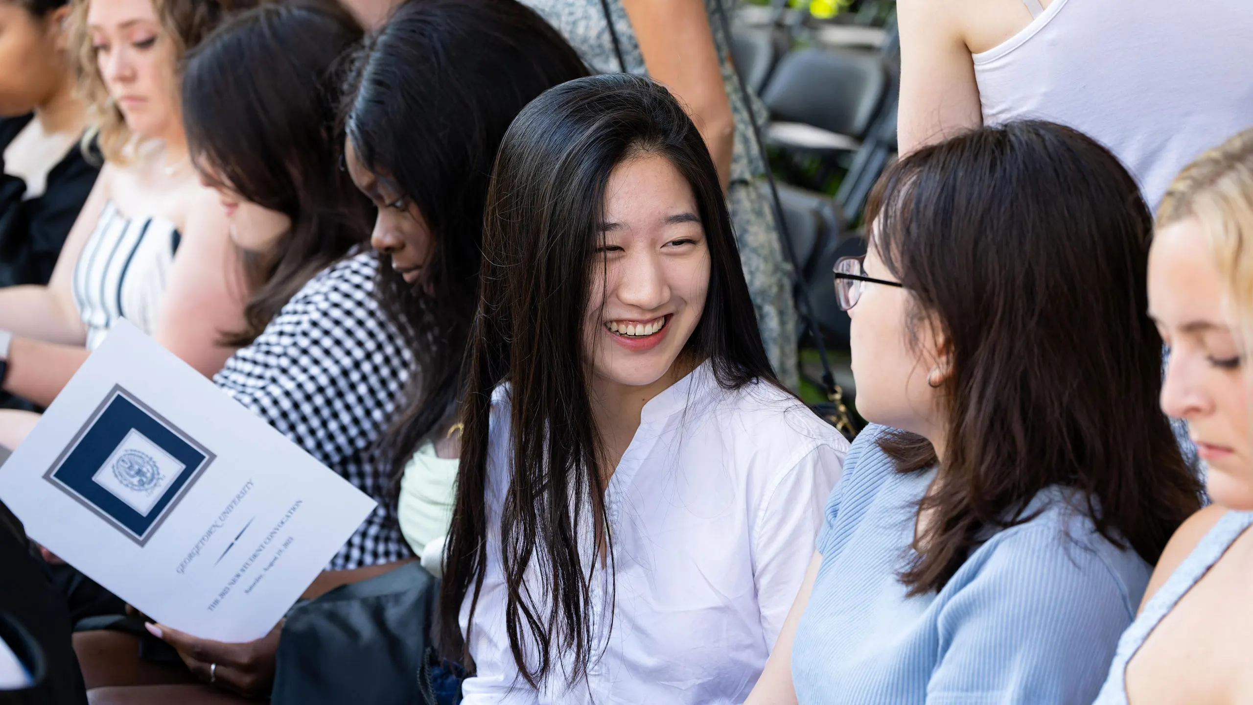 7 students sitting in a row at the new student orientation talking to each other, smiling.