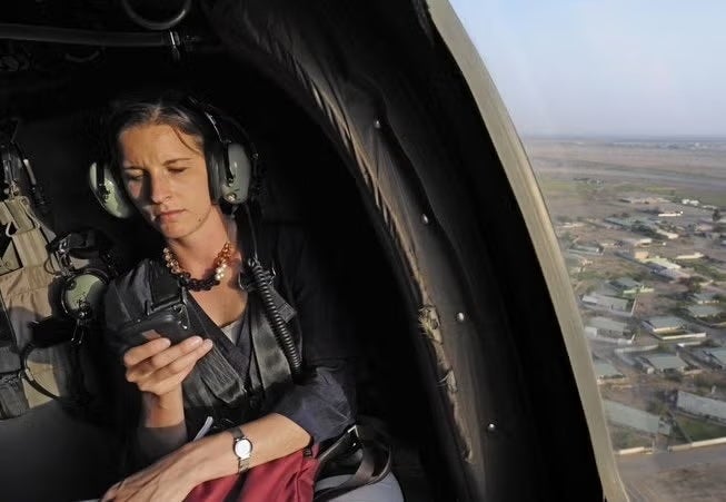 A person wearing headphones is using a smartphone inside a helicopter, looking focused, with a landscape visible through the window.