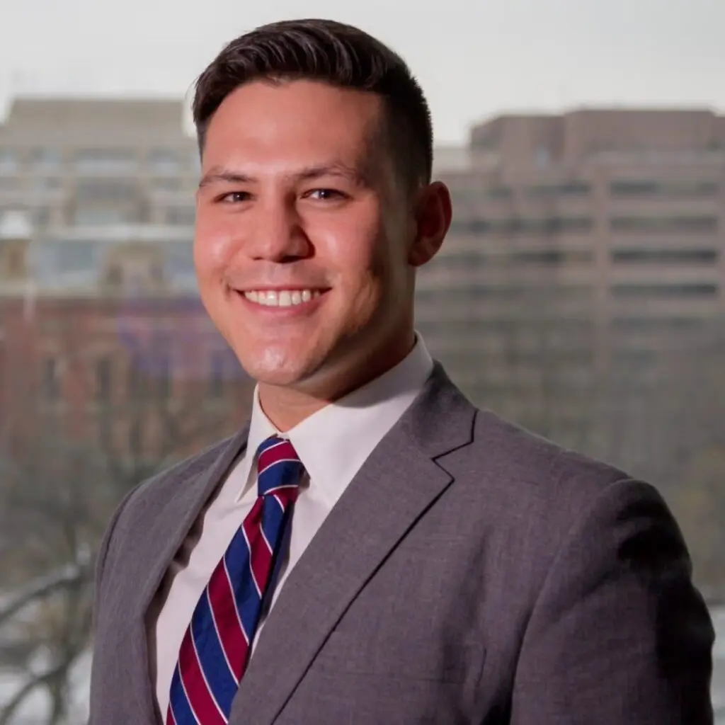 Benjamin Rich wearing a gray blazer, white, button-down shirt, and blue and red striped tie.