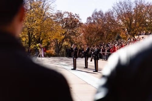 A ceremony at Arlington National Cemetery with uniformed honor guards and spectators in the background observing a wreath-laying ritual.