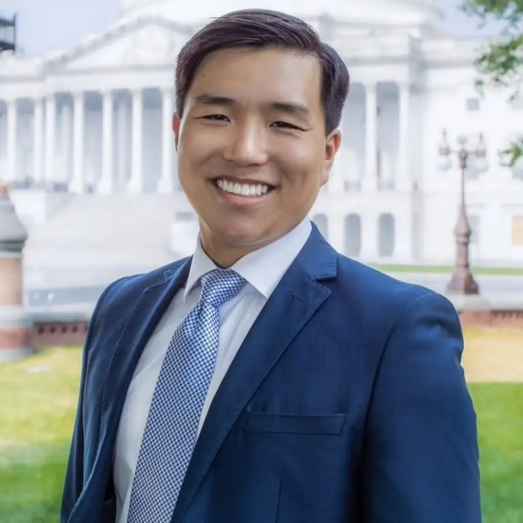 Christopher Cao wearing a blue blazer, white button-down shirt and a blue checkered tie a building with columns is in the background.