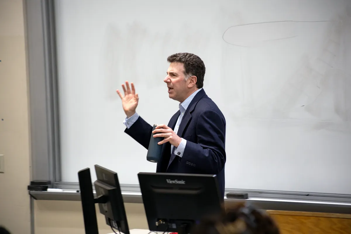 Georgetown professor wearing a suit speaking in front of a classroom. Behind him a clear whiteboard