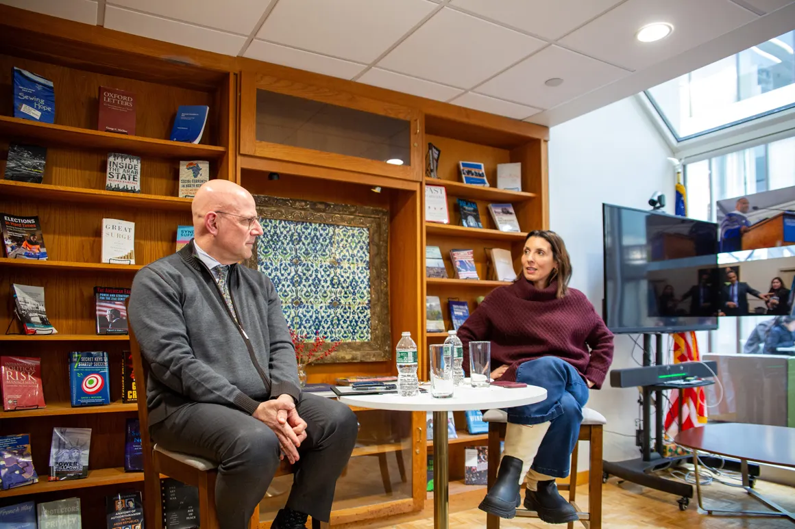 Dean Hellman and Lulu García Navarro in conversation, sitting opposite each other around a small table. Behind them a large bookshelf filled with books.