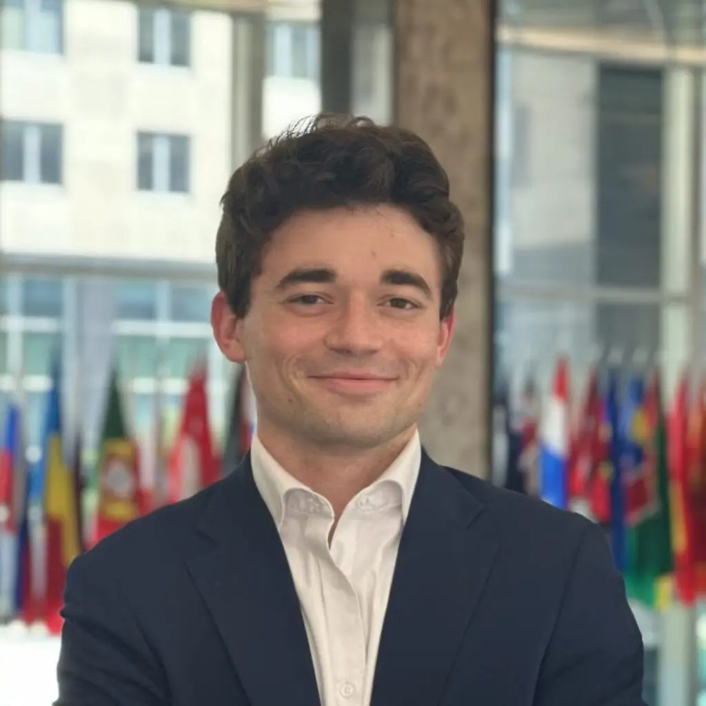 Collin Frank wearing a navy blue blazer and white button-down shirt with many flags in the background.