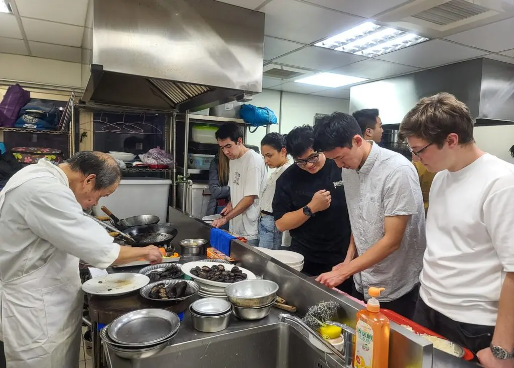 Five students in a kitchen observe an older man dressed as a chef working with mussels next to a sink
