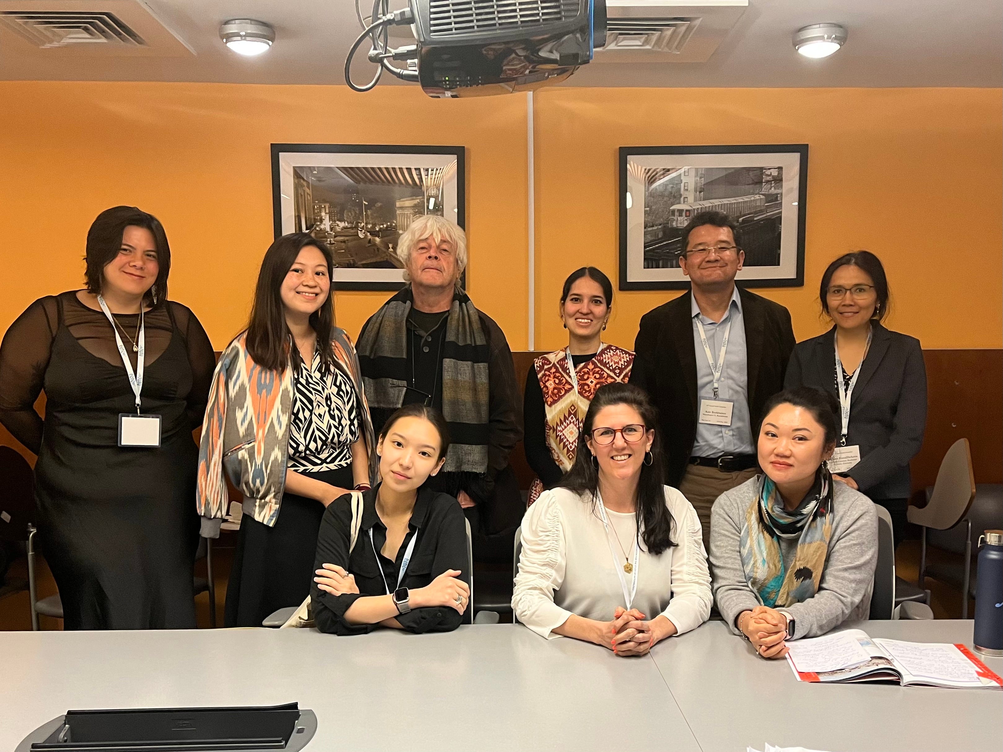 Darina Zhunussova sits at a table next to two women with six people standing behind her. They are wearing lanyards.