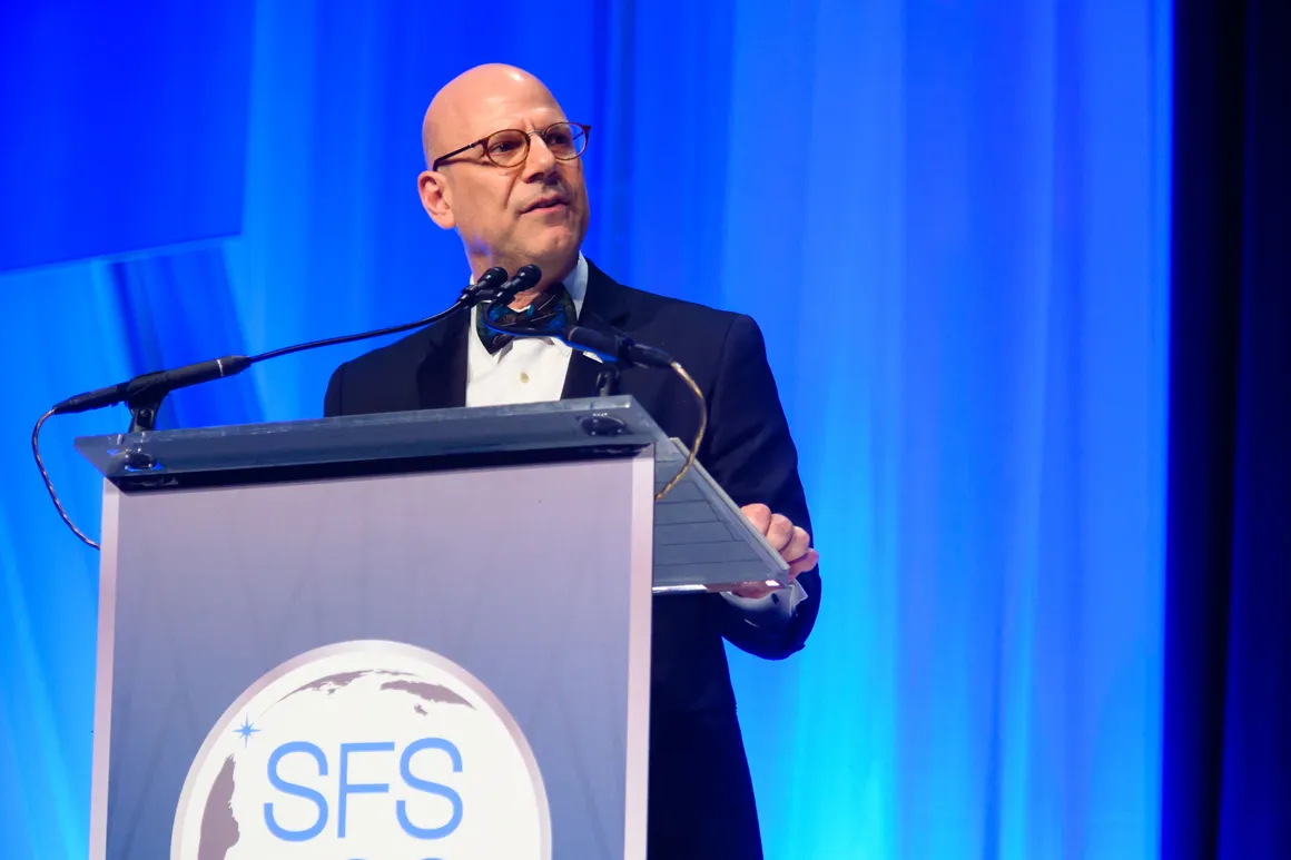 Dean Hellman dressed in formal attire, speaking on stage on a podium. The podium has the letters SFS on it.