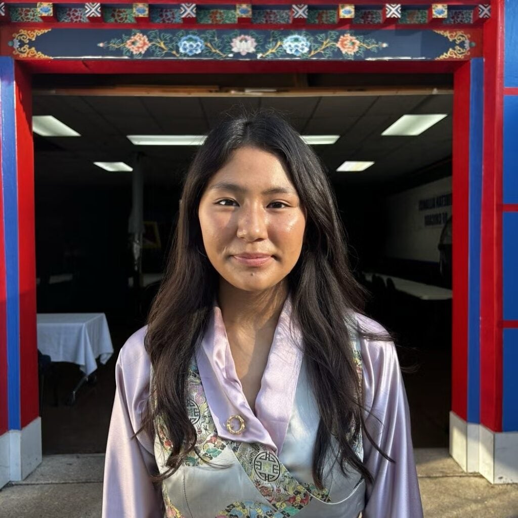 Dechen Atsatsang wearing traditional attire with a blue and red arch with floral designs in the background.