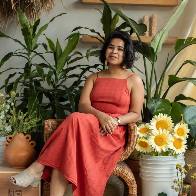 Devika Ranjan wearing a long orange dress, sitting on a rattan chair in front of green house plants and a bucket of bright yellow flours.