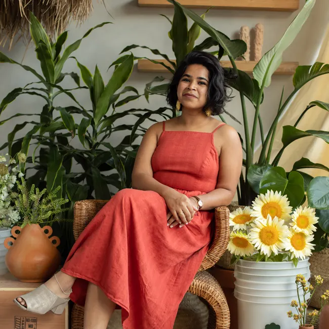 Devika Ranjan wearing a long orange dress, sitting on a rattan chair in front of green house plants and a bucket of bright yellow flours.