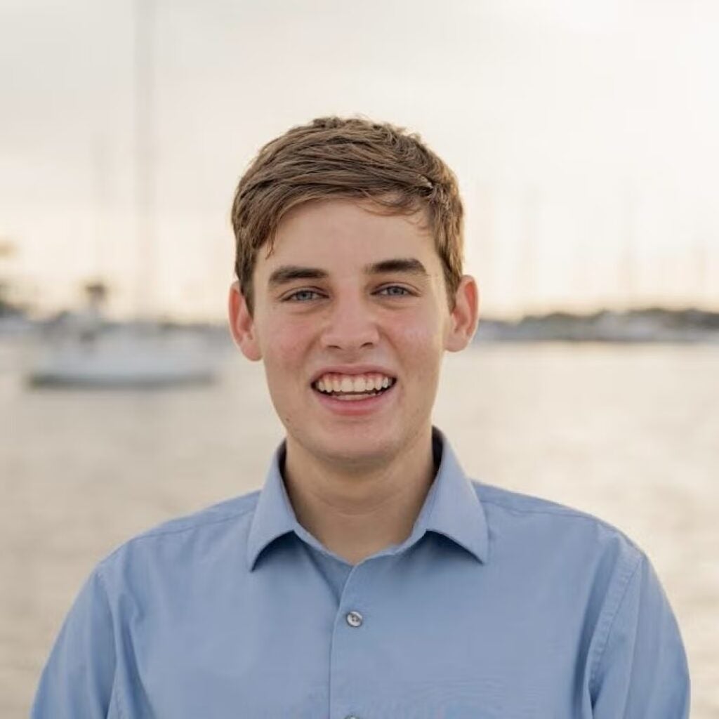Drew Zacharias wearing a light blue button-down shirt with blurred sailboats in the background.