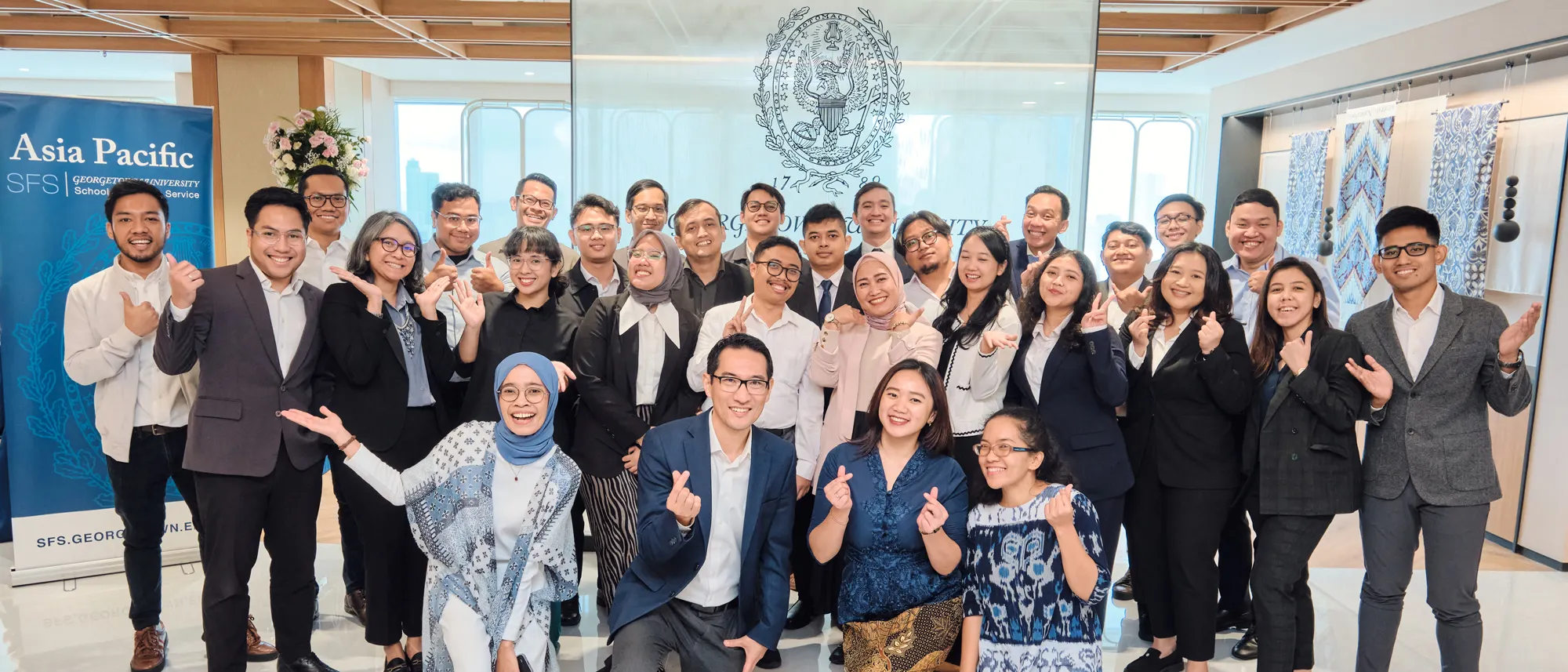 Group of students and faculty members posing for a group shot at the Georgetown SFS Asia Pacific in front of a window display with the Georgetown seal.