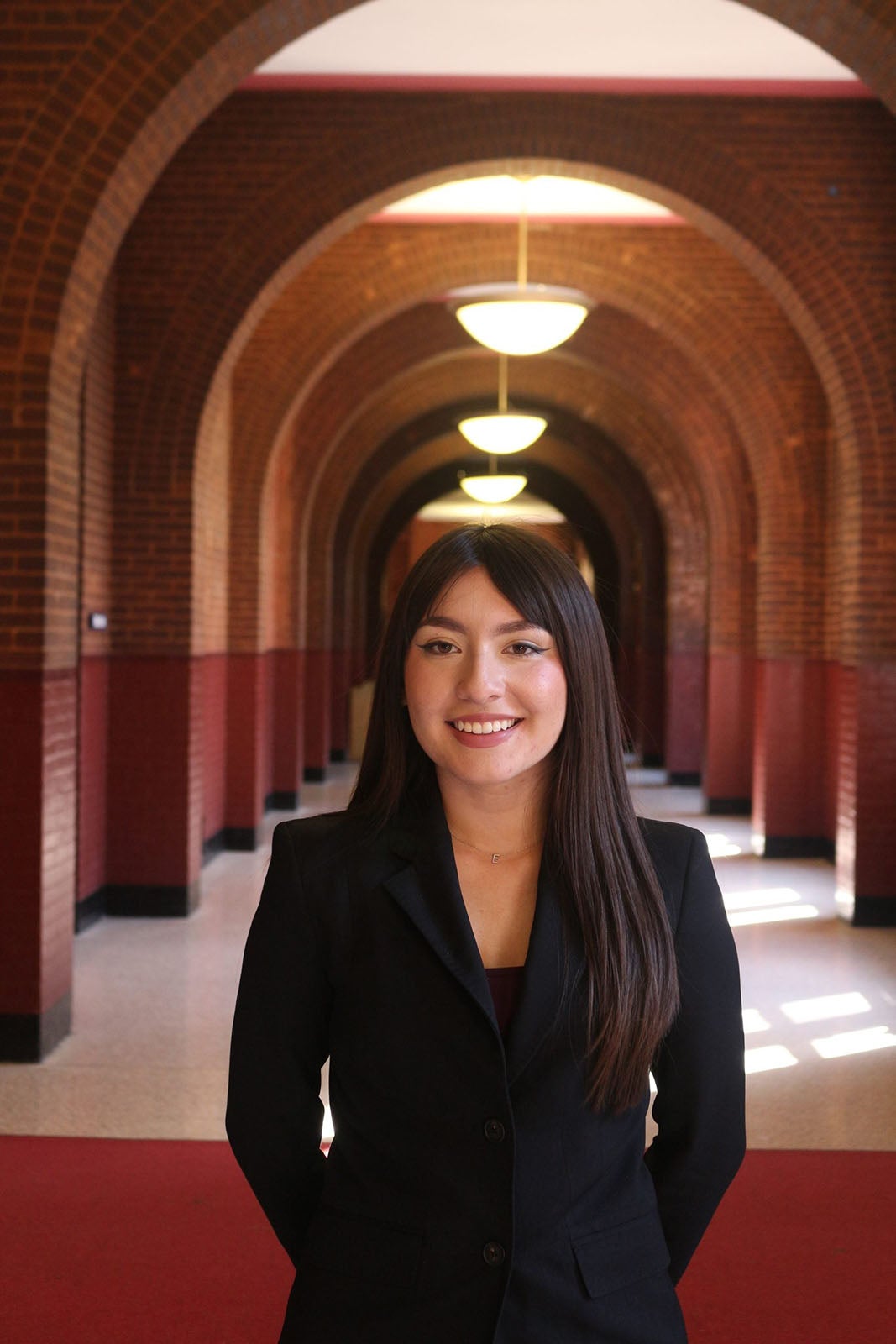 A young woman with long, straight, dark brown hair wearing a black suit and standing in a long hallway.