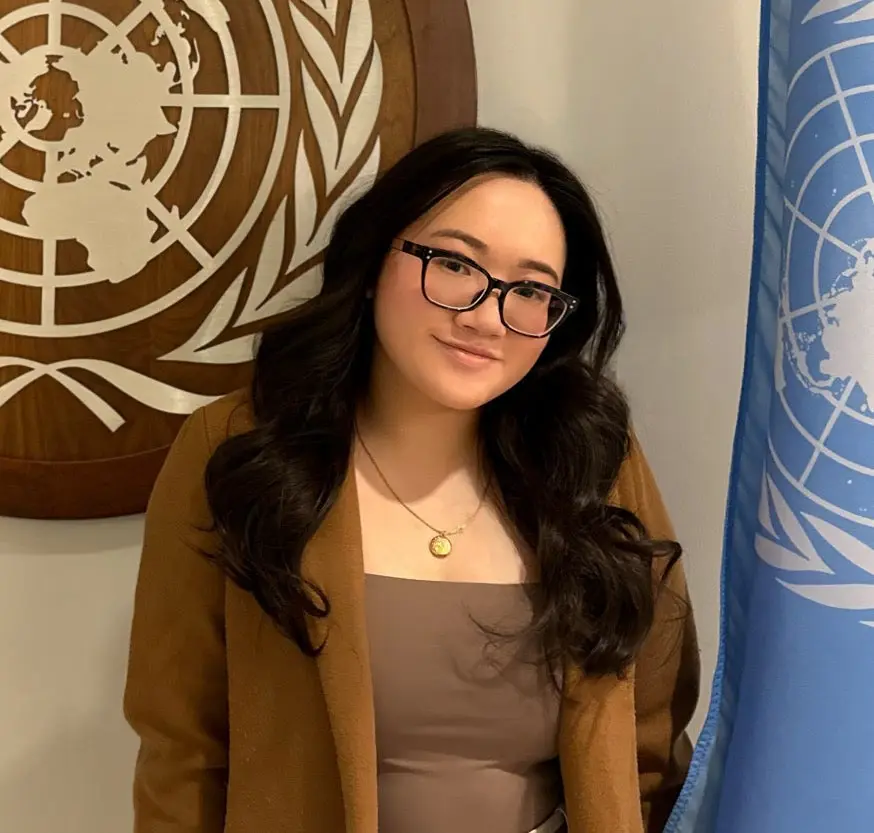 A young woman with glasses and long dark hair wearing a camel colored suit jacket and a brown short with a gold locket.