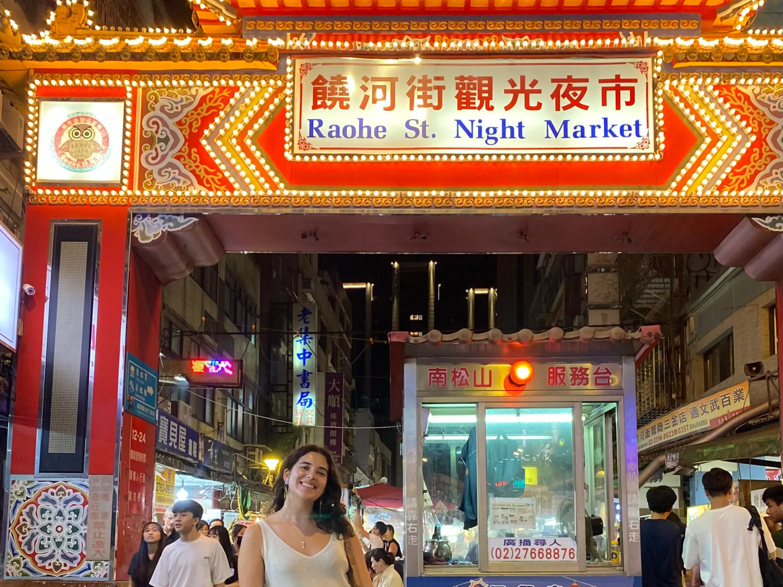 Person smiling in front of the brightly lit entrance to Raohe Street Night Market in Taiwan, surrounded by bustling street vendors and visitors.